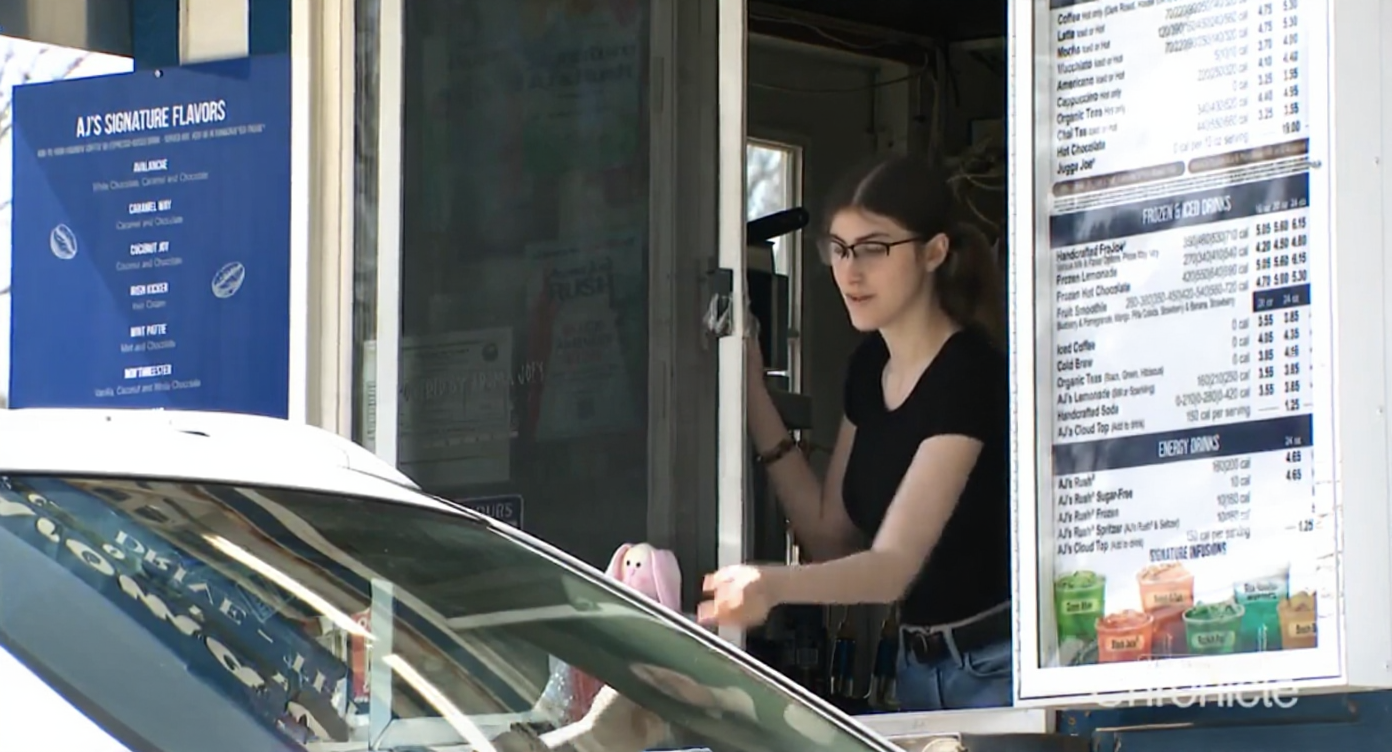 A picture of a girl with glasses on handing coffee out to a drive-through customer.